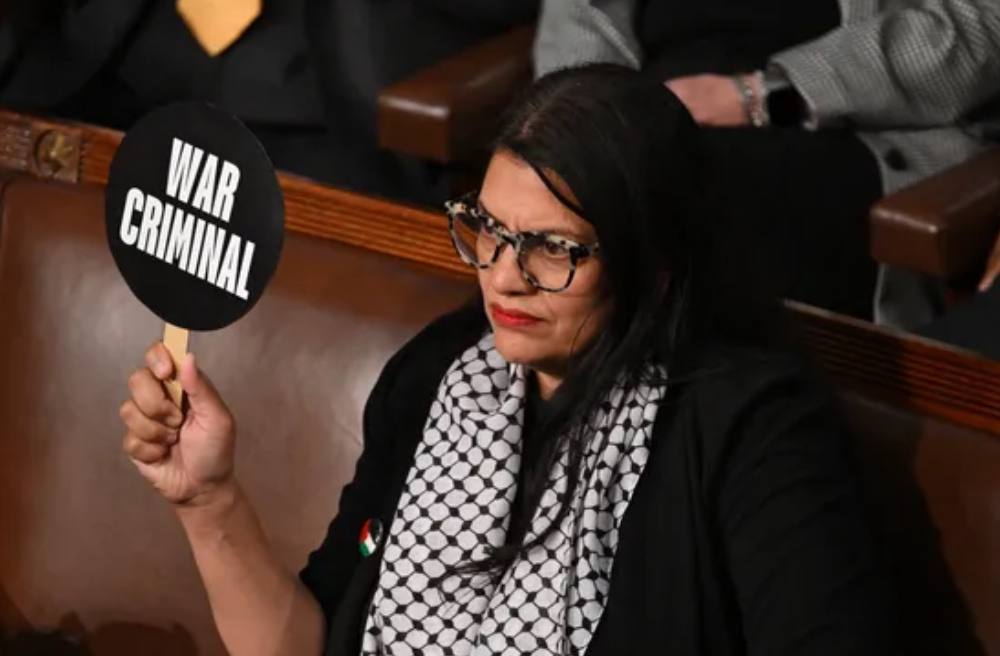 La députée américaine Rashida Tlaib (DSA) tient un panneau sur lequel on peut lire  » criminel de guerre  » (et au verso  » coupable de genocide « ) lors du discours du Premier ministre israélien au Congrès.
